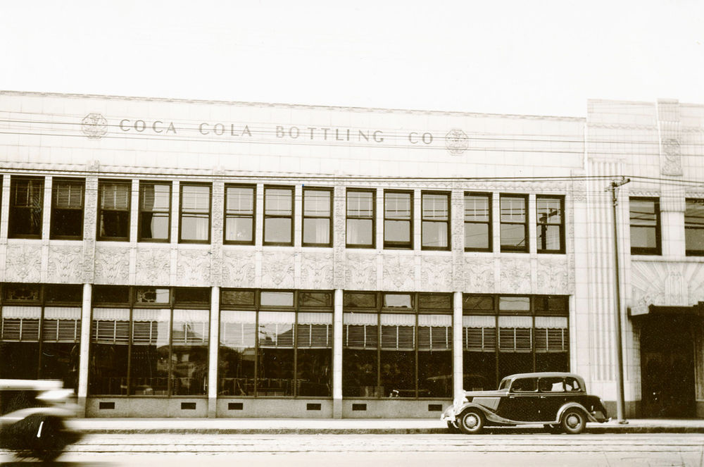 Archival photograph of the Coca-Cola bottling plant façade with vintage cars parked outside.